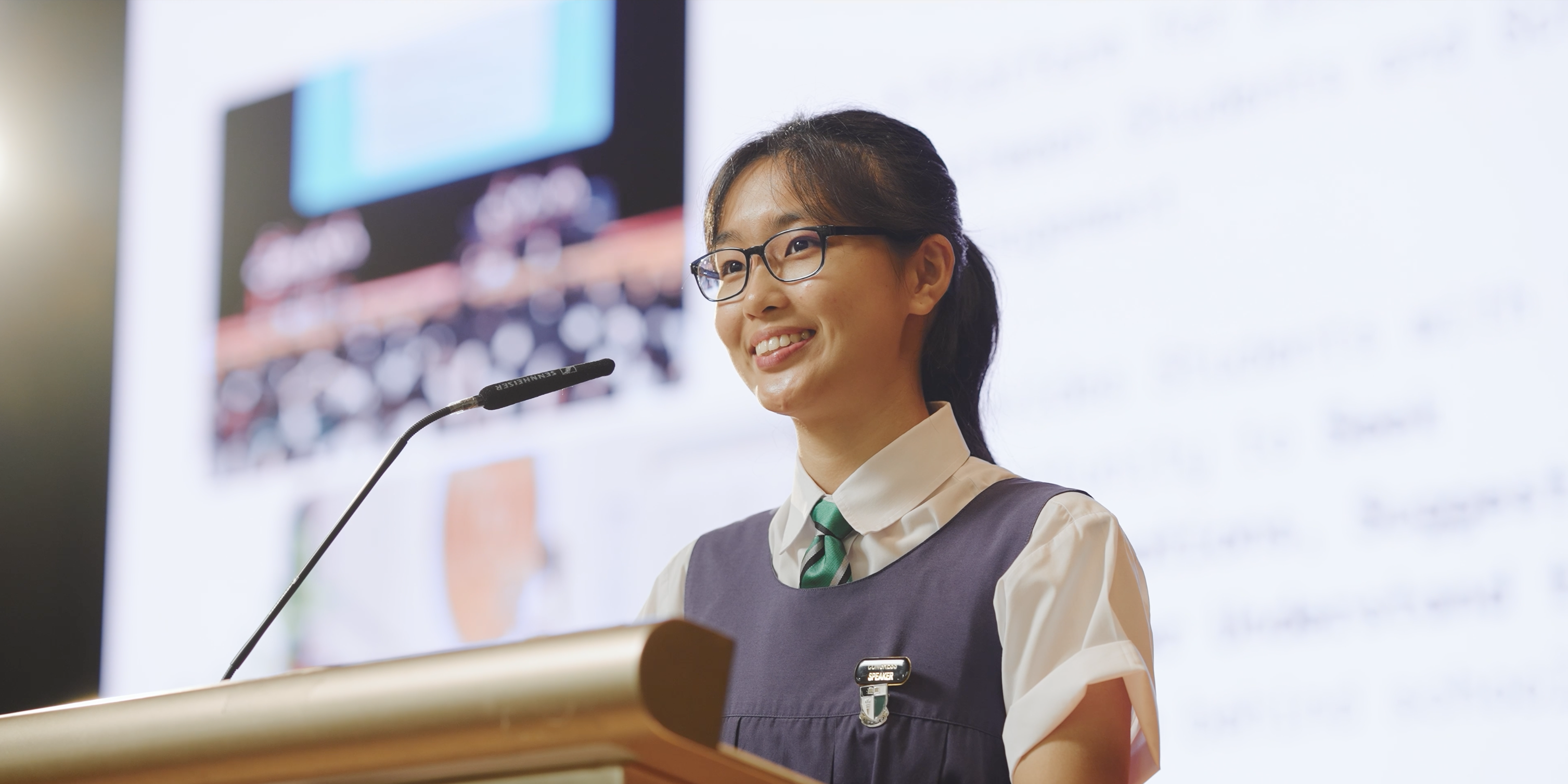 A female student smiling while standing behind a microphone and rostrum, with a projected screen behind.